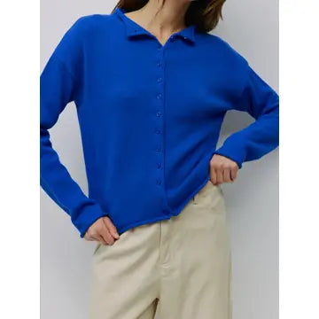 A woman wearing a bright royal blue long-sleeve button-front collared top with beige pants, cropped from the neck down against a plain light gray background.