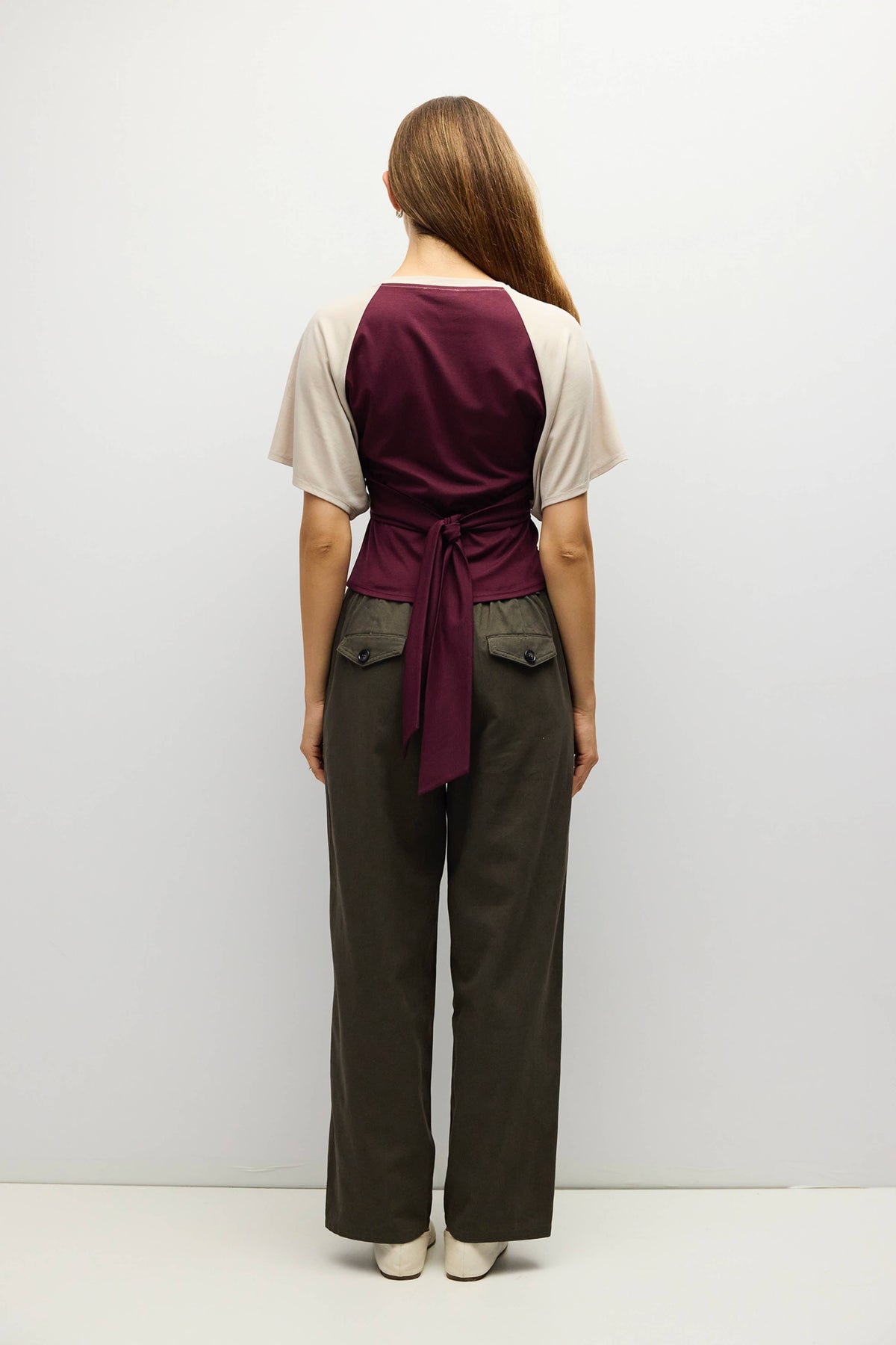 A woman viewed from the back wearing a short-sleeve wrap-style top in deep burgundy and cream, paired with dark olive wide-leg pants and white shoes, against a plain light gray studio background.