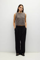 A woman stands against a plain light gray wall wearing a sleeveless textured knit top in heathered taupe-brown and black wide-leg trousers, with black flats.