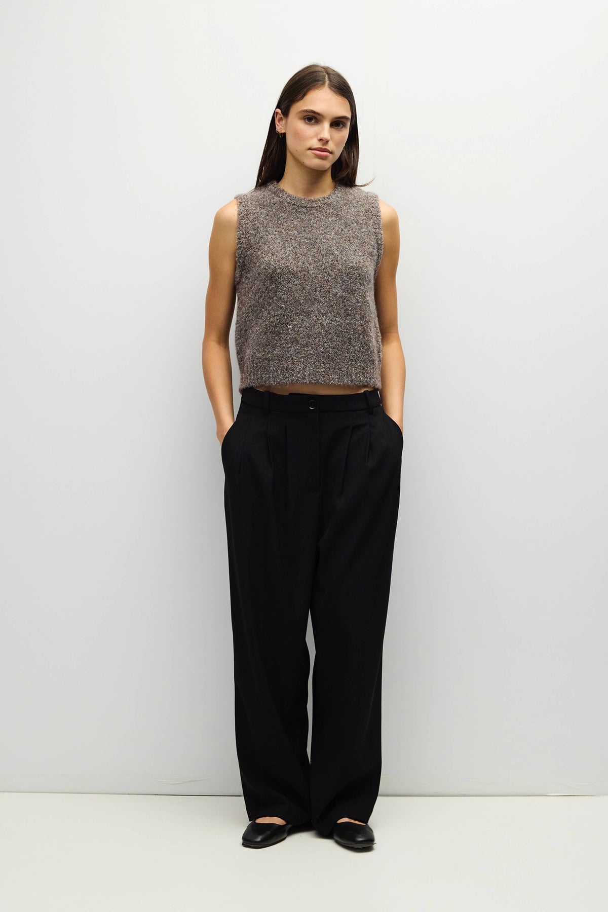 A woman stands against a plain light gray wall wearing a sleeveless textured knit top in heathered taupe-brown and black wide-leg trousers, with black flats.