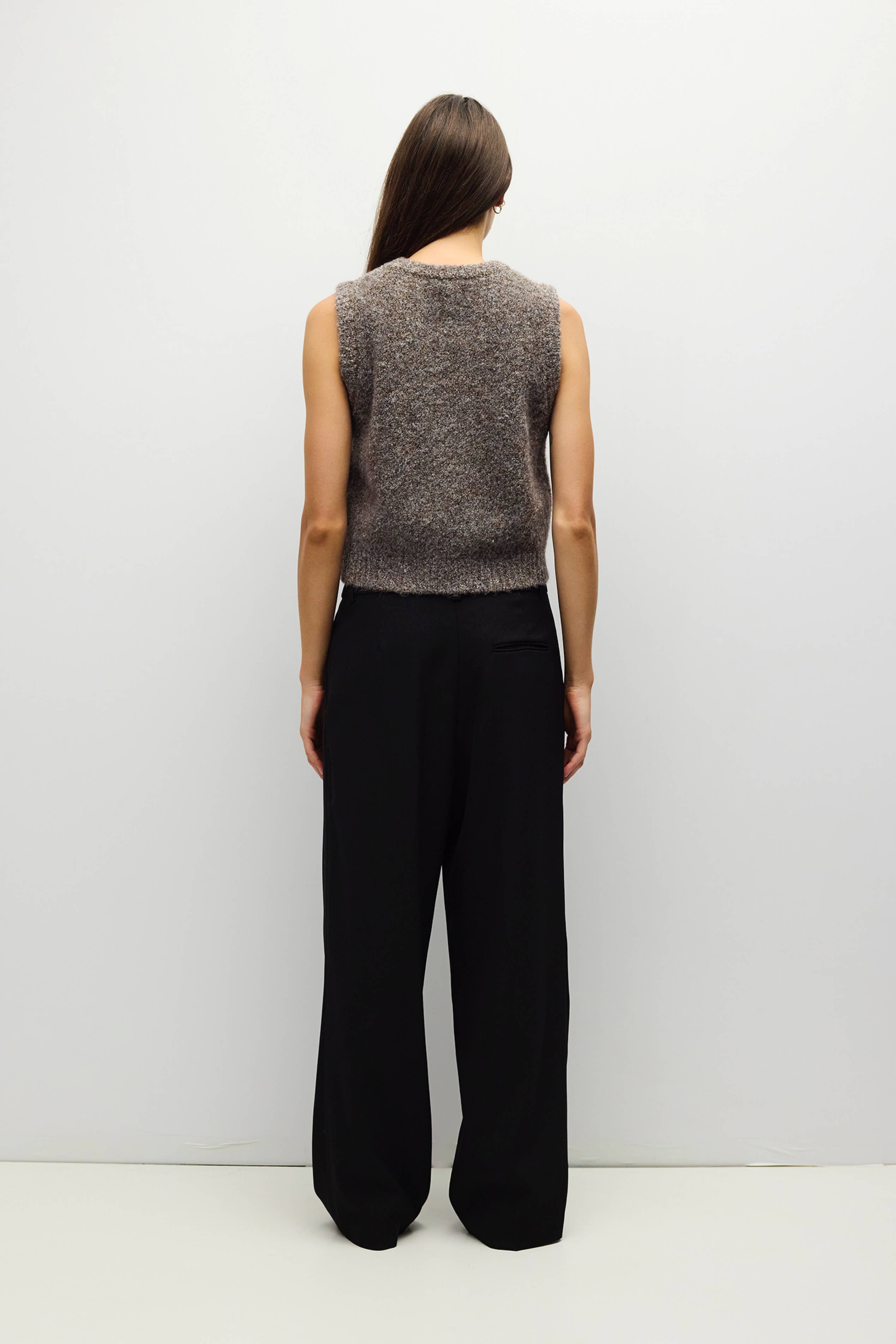 A woman viewed from the back wearing a sleeveless marled gray knit top and black wide-leg trousers, standing against a plain light gray studio background.