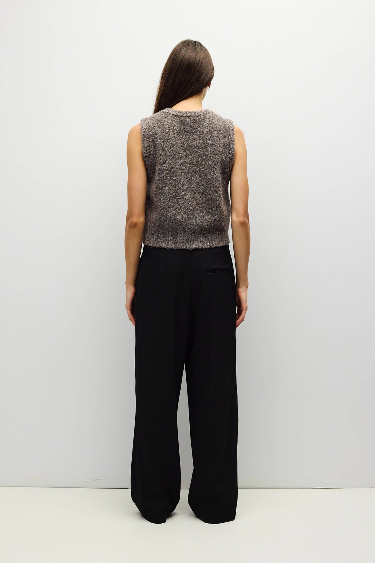 A woman viewed from the back wearing a sleeveless marled gray knit top and black wide-leg trousers, standing against a plain light gray studio background.