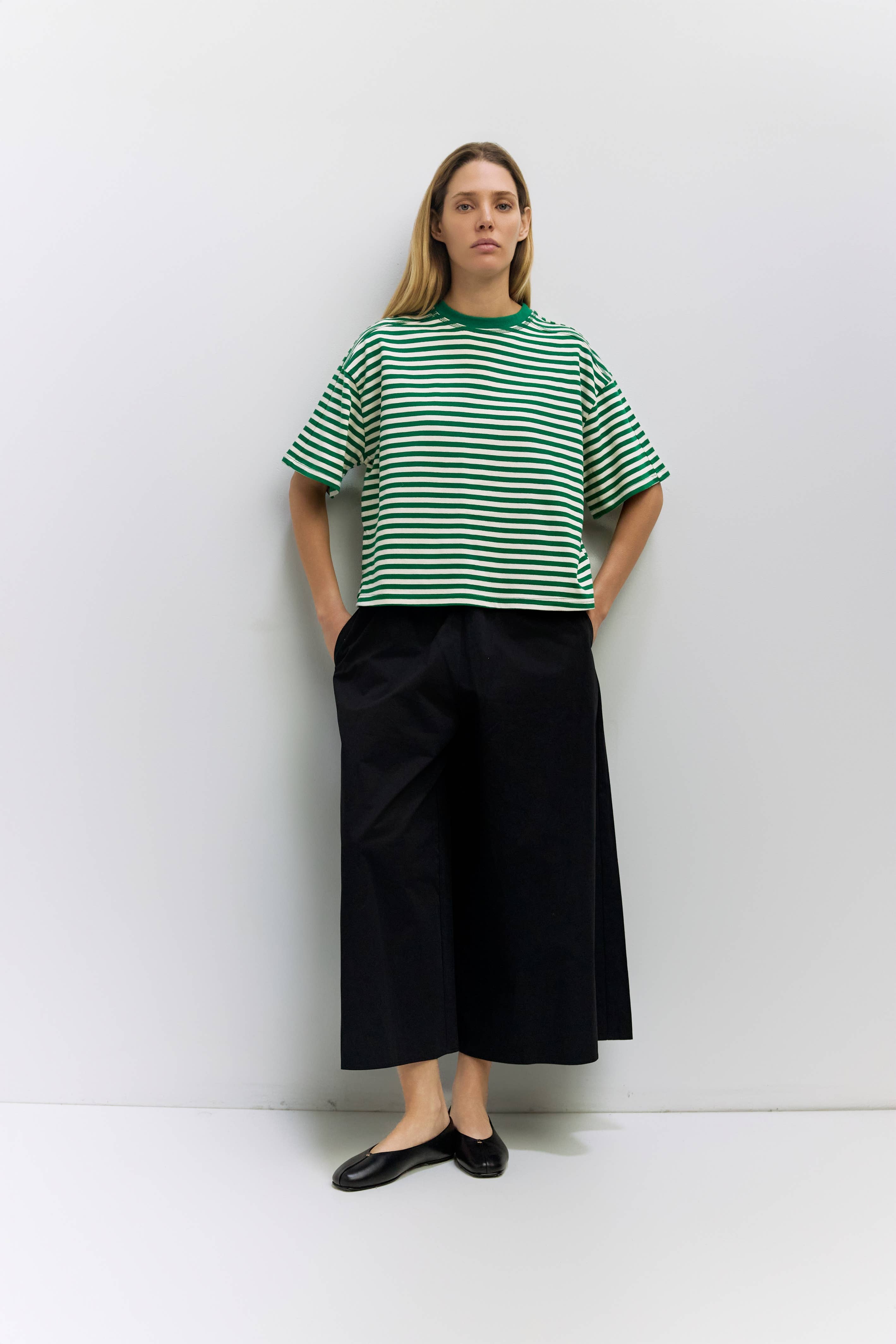 A woman stands against a light gray wall wearing an oversized green and white striped short-sleeve T-shirt, black wide-leg cropped pants, and black flats.