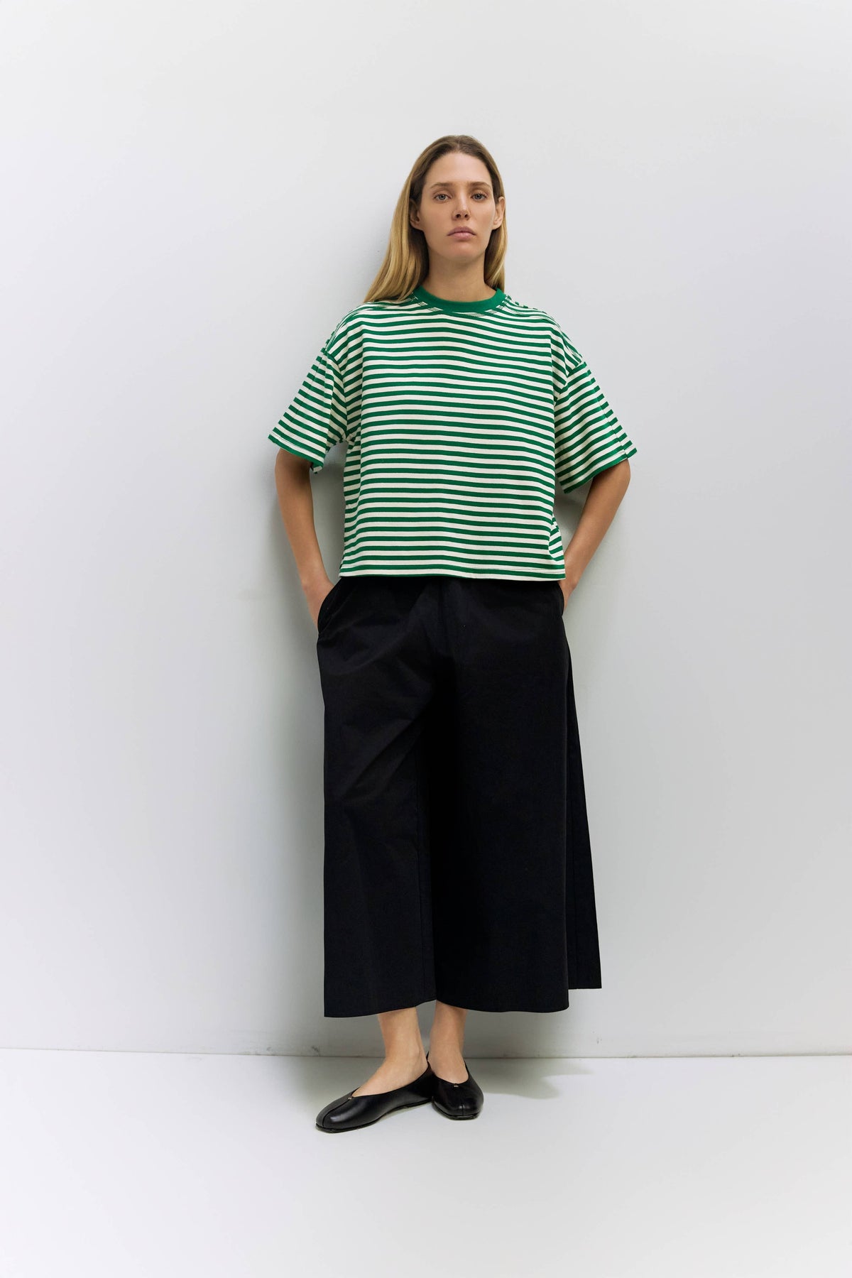 A woman stands against a light gray wall wearing an oversized green and white striped short-sleeve T-shirt, black wide-leg cropped pants, and black flats.