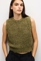 A woman wearing a sleeveless, crew-neck knit sweater vest in a heathered olive green and brown color, paired with black trousers, against a plain light gray background.