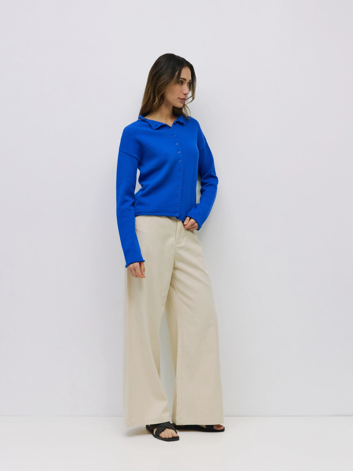 A woman modeling a bright blue long-sleeve button-up shirt paired with wide-leg beige trousers and black flat sandals, standing against a plain white studio background.