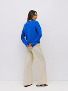 A woman stands with her back to the camera wearing a bright blue cropped long-sleeve top and wide-leg beige trousers, paired with black sandals, against a plain white studio background.