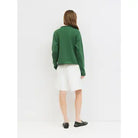 A woman viewed from behind wearing a dark green long-sleeve cropped sweater, a white skirt, and black loafers, standing against a plain white studio background.