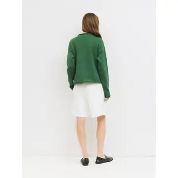 A woman viewed from behind wearing a dark green long-sleeve cropped sweater, a white skirt, and black loafers, standing against a plain white studio background.