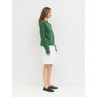 A woman stands in profile wearing a green cropped knit cardigan, a white knee-length skirt, and black loafers against a plain white studio background.