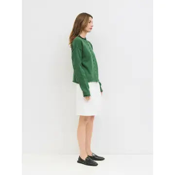 A woman stands in profile wearing a green cropped knit cardigan, a white knee-length skirt, and black loafers against a plain white studio background.