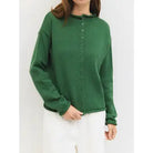 A woman wearing a green ribbed knit long-sleeve button-up sweater and white pants, shown from the neck down against a plain light background.