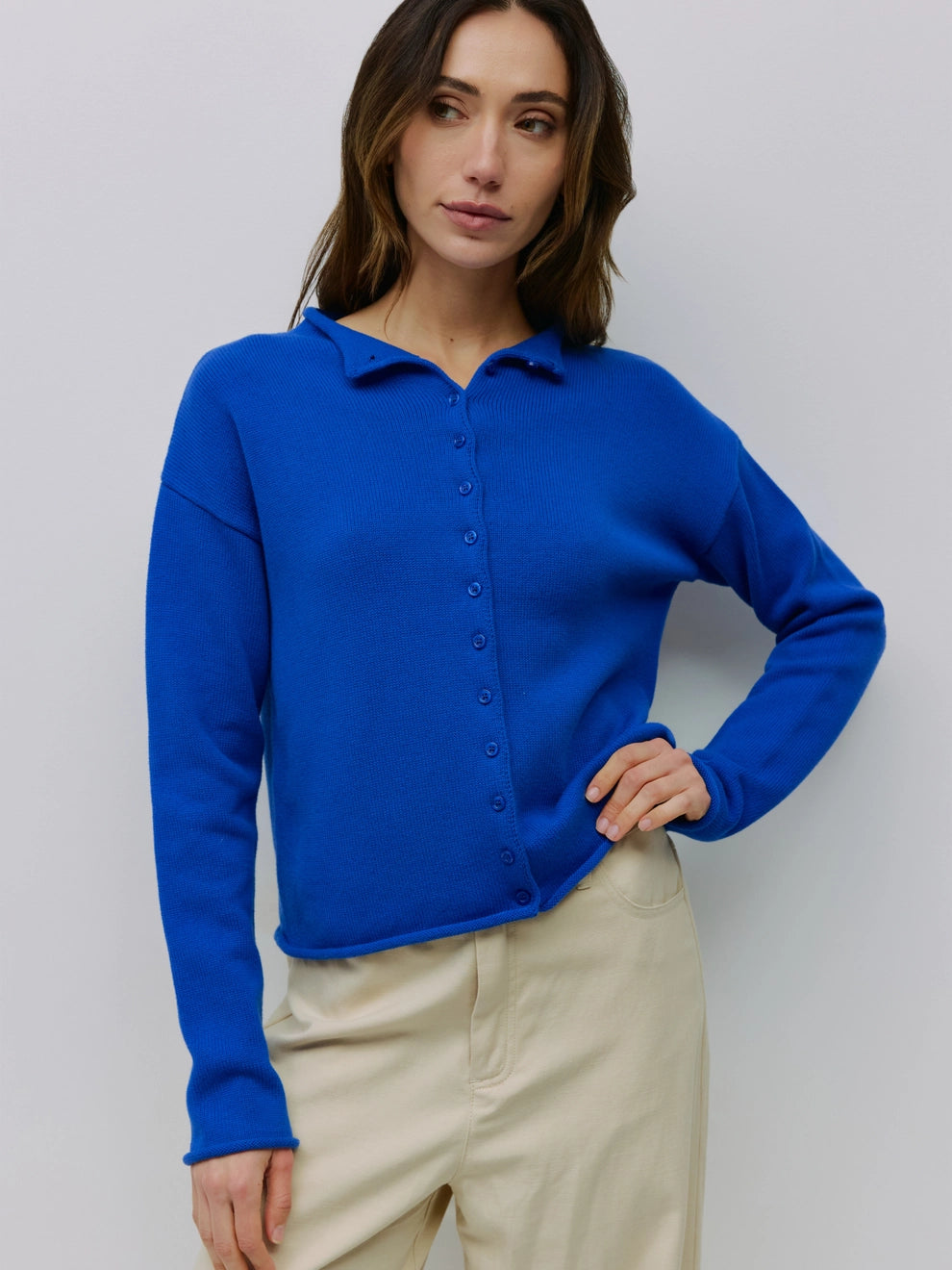 A woman wearing a bright royal blue long-sleeve knit polo-style top with a button placket, paired with light beige pants, standing against a plain light gray studio background.