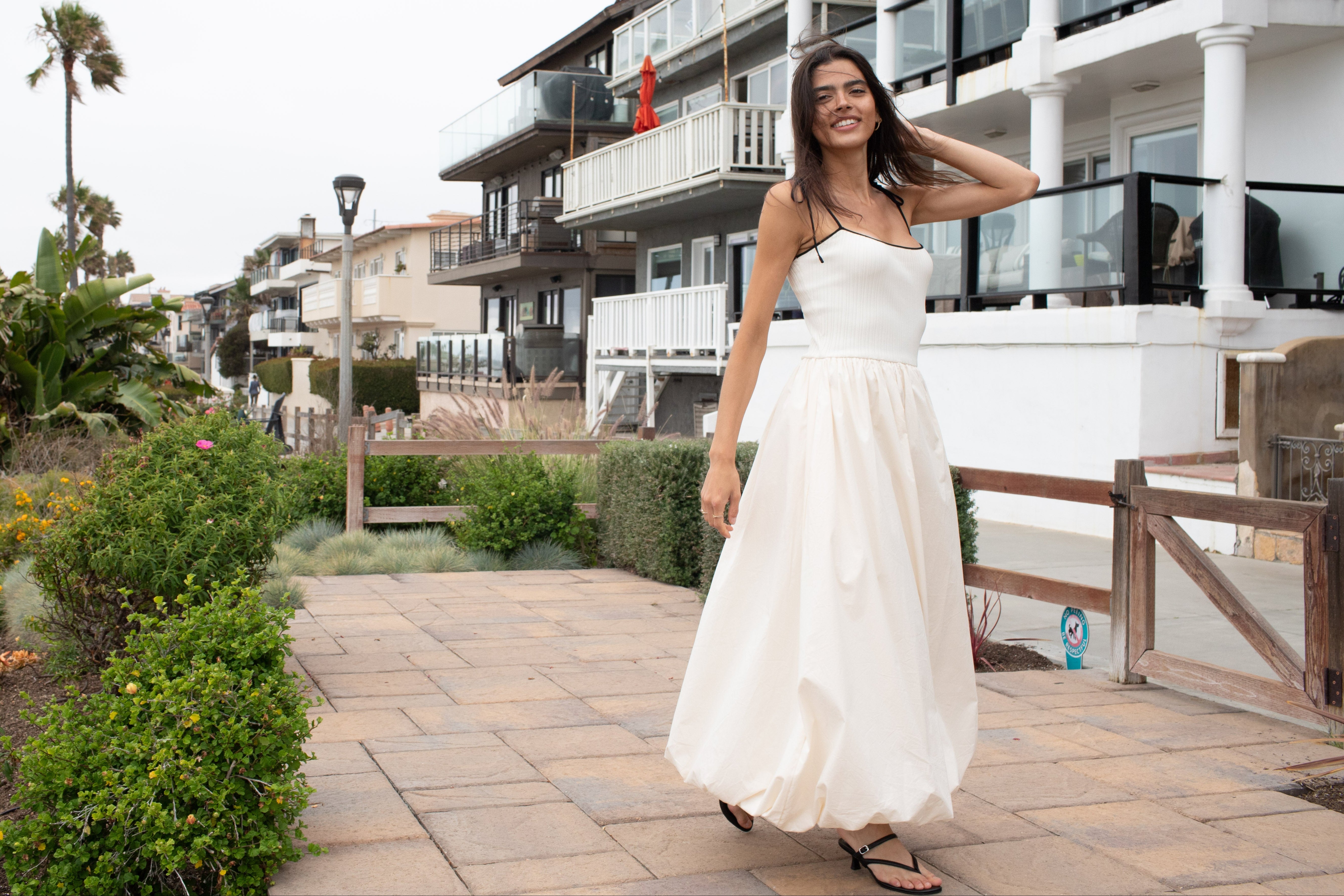 Woman in an ivory dress with bubble hem and shoulder ties standing on a boardwalk with beach houses and palm trees in the background.