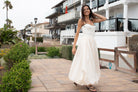 Woman in an ivory dress with bubble hem and shoulder ties standing on a boardwalk with beach houses and palm trees in the background.