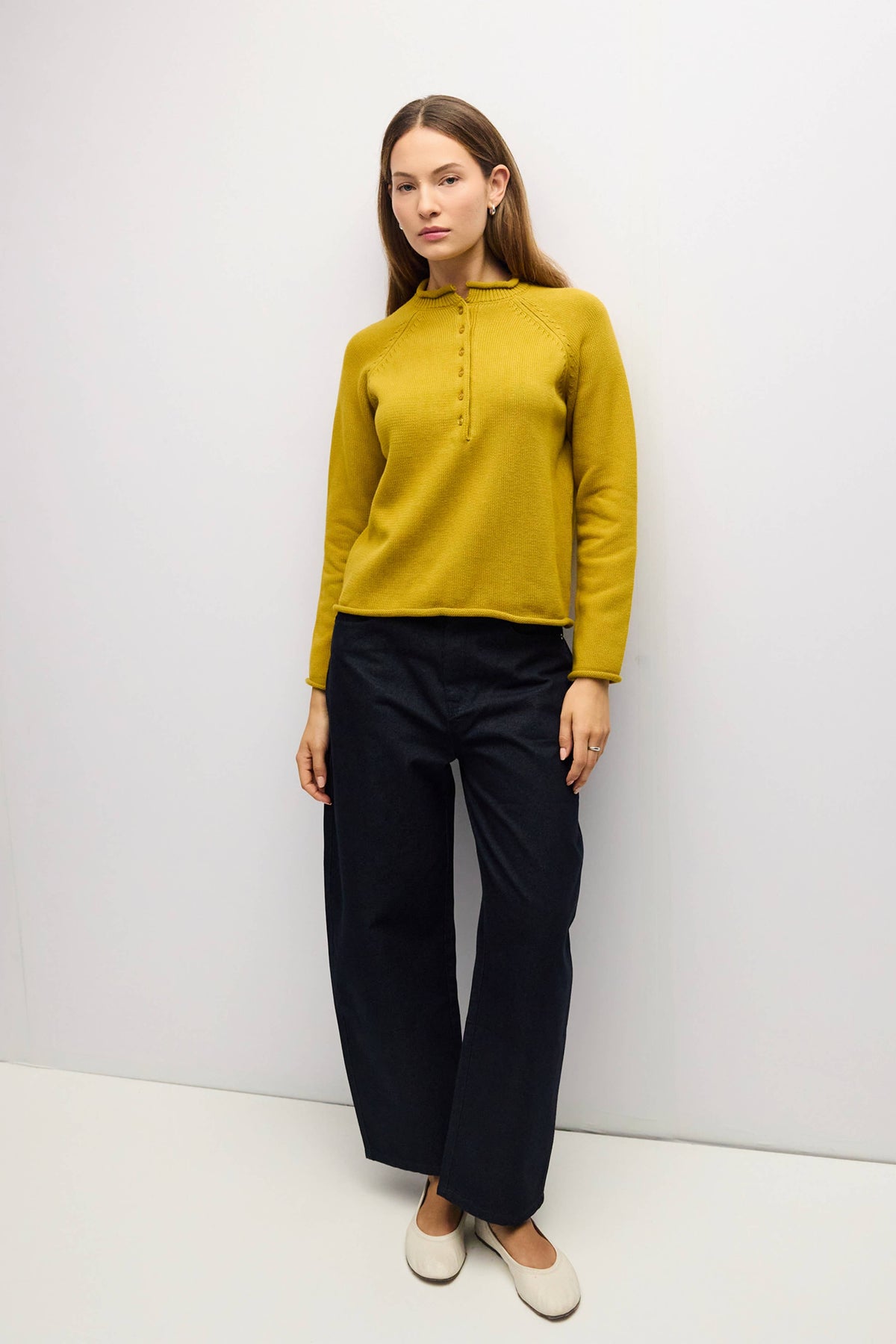 A woman modeling a mustard yellow mock-neck knit top with a button placket, paired with dark navy wide-leg trousers and off-white flat shoes, standing against a plain white studio background.