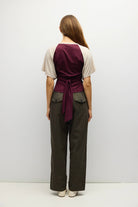 A woman viewed from the back wearing a short-sleeve wrap-style top in deep burgundy and cream, paired with dark olive wide-leg pants and white shoes, against a plain light gray studio background.