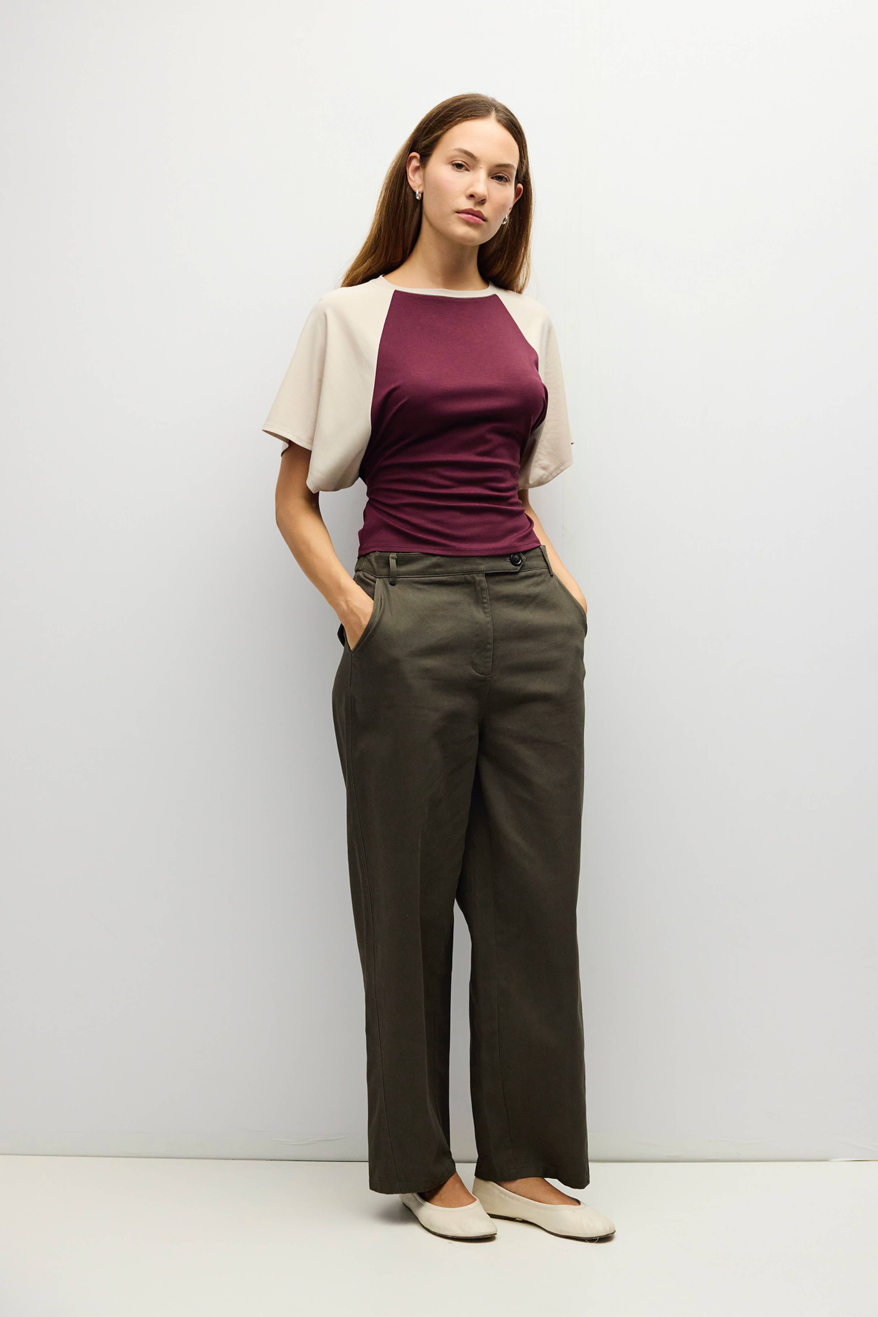A woman modeling a short-sleeve raglan top with cream sleeves and a deep burgundy body, paired with dark olive wide-leg trousers and beige flats, standing against a light gray studio background.