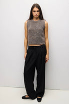A woman stands against a plain light gray studio wall wearing a sleeveless textured knit top in heathered gray-brown tones, loose black wide-leg pants, and black flats.