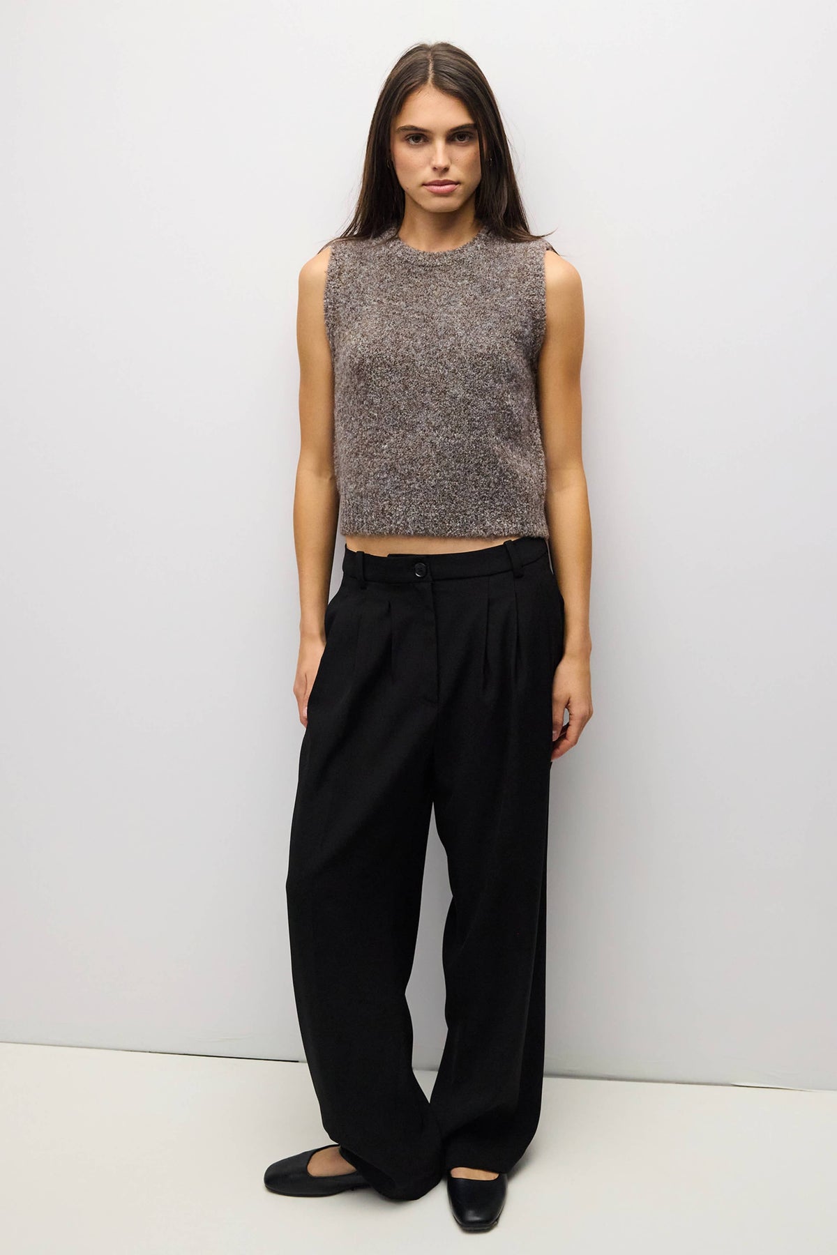 A woman stands against a plain light gray studio wall wearing a sleeveless textured knit top in heathered gray-brown tones, loose black wide-leg pants, and black flats.
